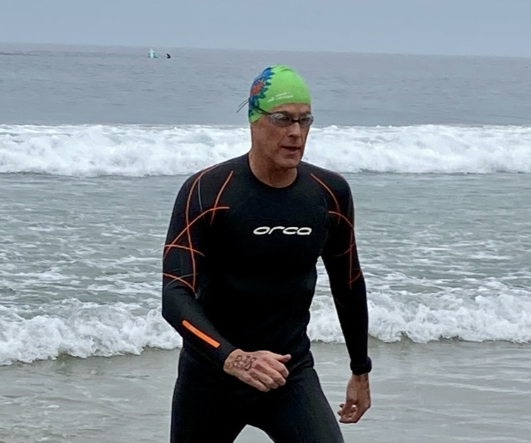 Crick Waters emerging from open-water swimming in La Jolla, wearing a wetsuit and swim cap